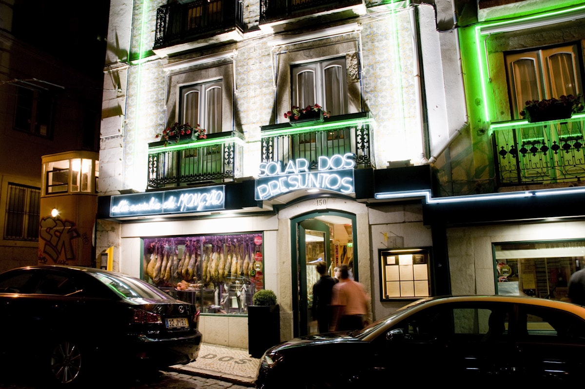 The neon-lit facade of Solar dos Presuntos, a Michelin-recommended restaurant in Rua das Portas de Santo Antpo noted for its fish and seafood choices. The Elevador do Lavra (funicular) is stationary to the left of the building.