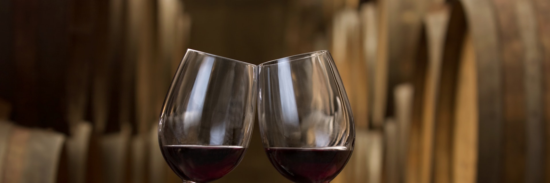 "Two hands holding wineglasses wi red wine make a toast in a cellar in front of a row of oak barrels,See also:"