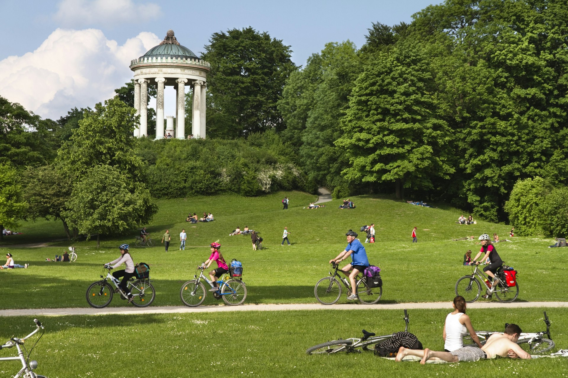Cyclists on a bike path pedal past sunbathers and picnickers on the lawns of the English Garden, Munich