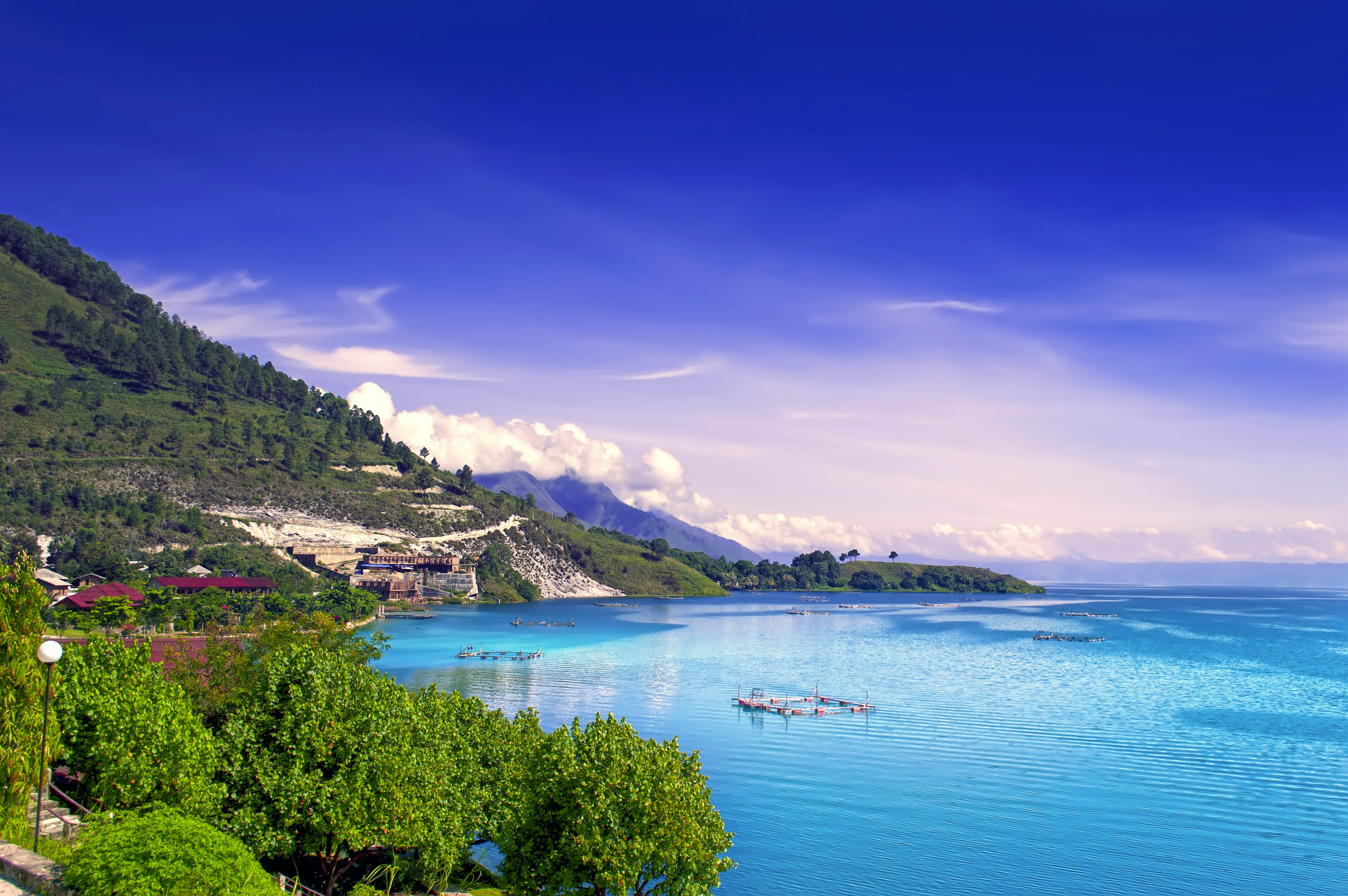  The image shows a beautiful view of Danau Toba, a large volcanic lake in Sumatra, Indonesia. The lake is surrounded by green hills and mountains, and the water is crystal clear.