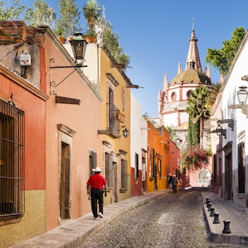 Man walking down a cobblestone street with chuch dome in distance.