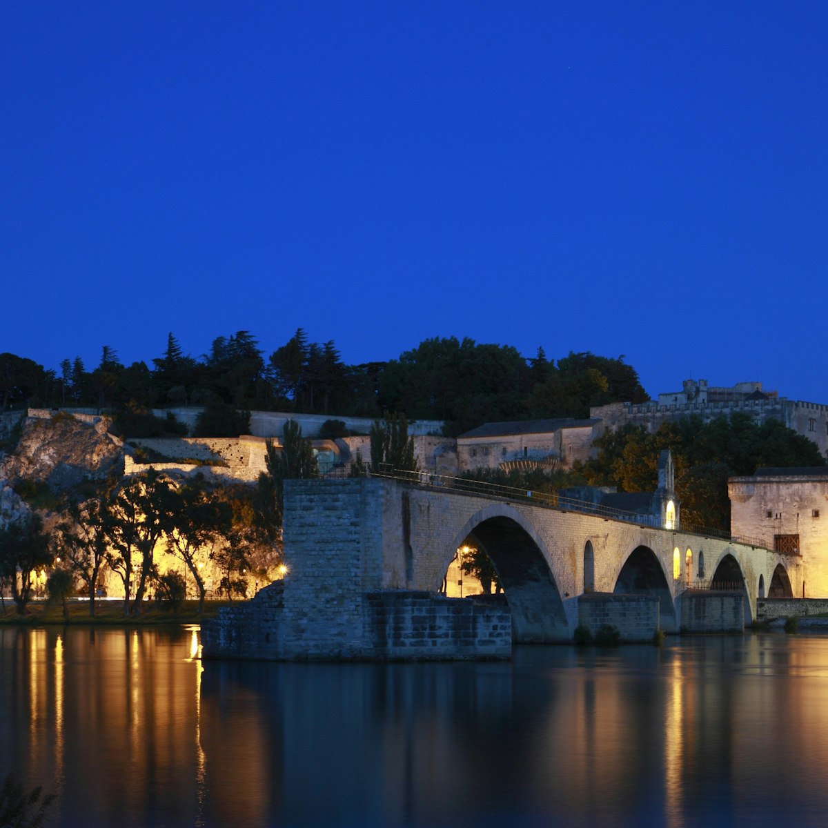 The Avignon Papal Palace (Palais des Papes) and the Avignon Bridge (Pont d'Avignon or Pont St-Bénézet) illuminated at night under the deep blue sky reflecting in the still waters of Rhone river. ; Shutterstock ID 609704714