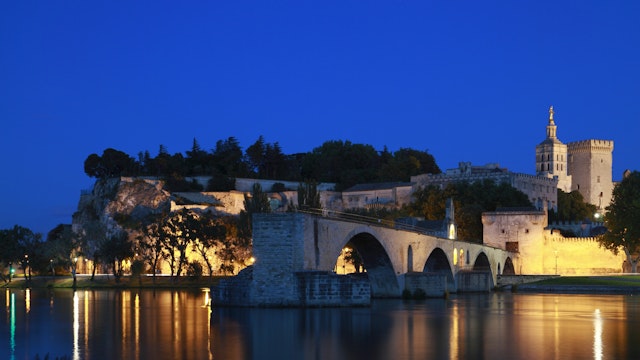 The Avignon Papal Palace (Palais des Papes) and the Avignon Bridge (Pont d'Avignon or Pont St-Bénézet) illuminated at night under the deep blue sky reflecting in the still waters of Rhone river. ; Shutterstock ID 609704714
