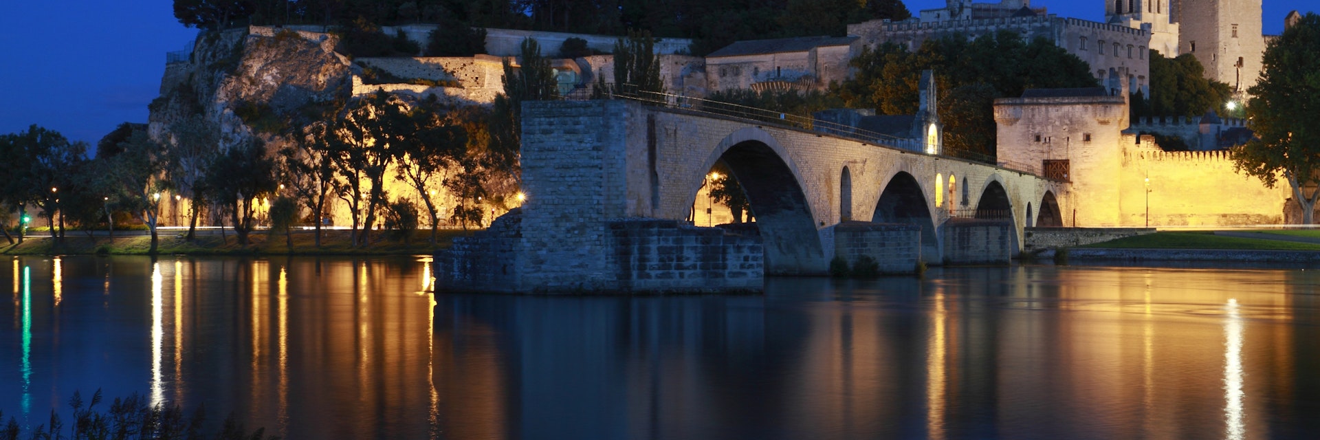 The Avignon Papal Palace (Palais des Papes) and the Avignon Bridge (Pont d'Avignon or Pont St-Bénézet) illuminated at night under the deep blue sky reflecting in the still waters of Rhone river. ; Shutterstock ID 609704714