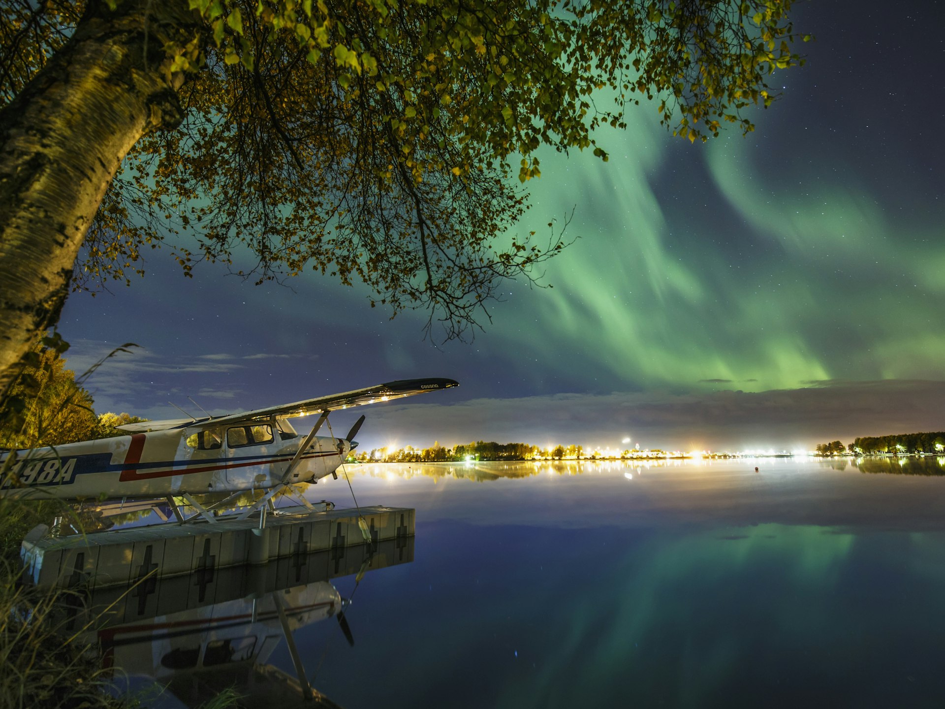 The green glow of the Northern Lights illuminates a seaplane, trees and houses by night at Lake Hood