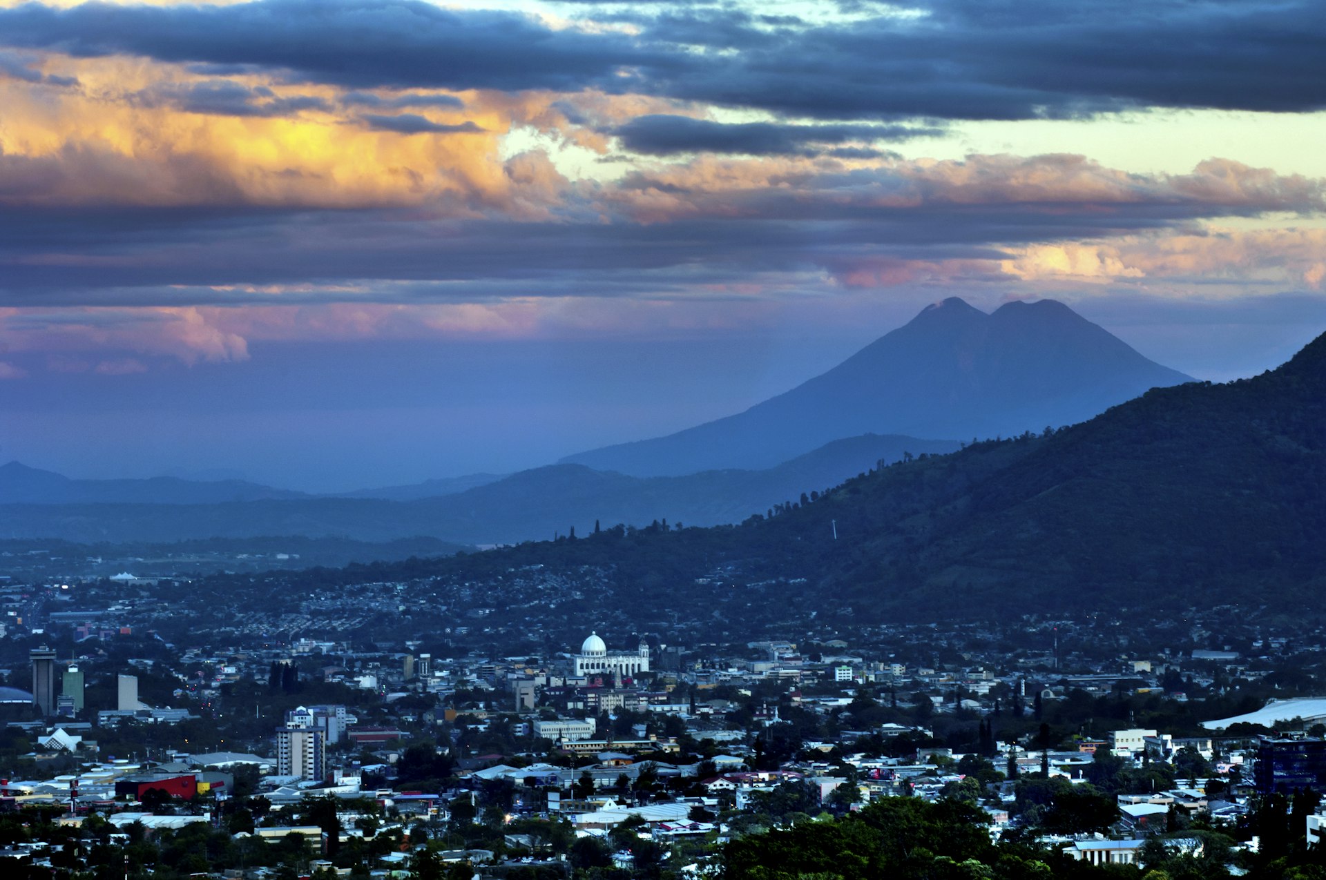 A wide sunset view of the skyline of San Salvador (including the white-domed Metropolitan Cathedral Of The Holy Savior) with the volcano El Boquerón’s peaks in the distance, El Salvador