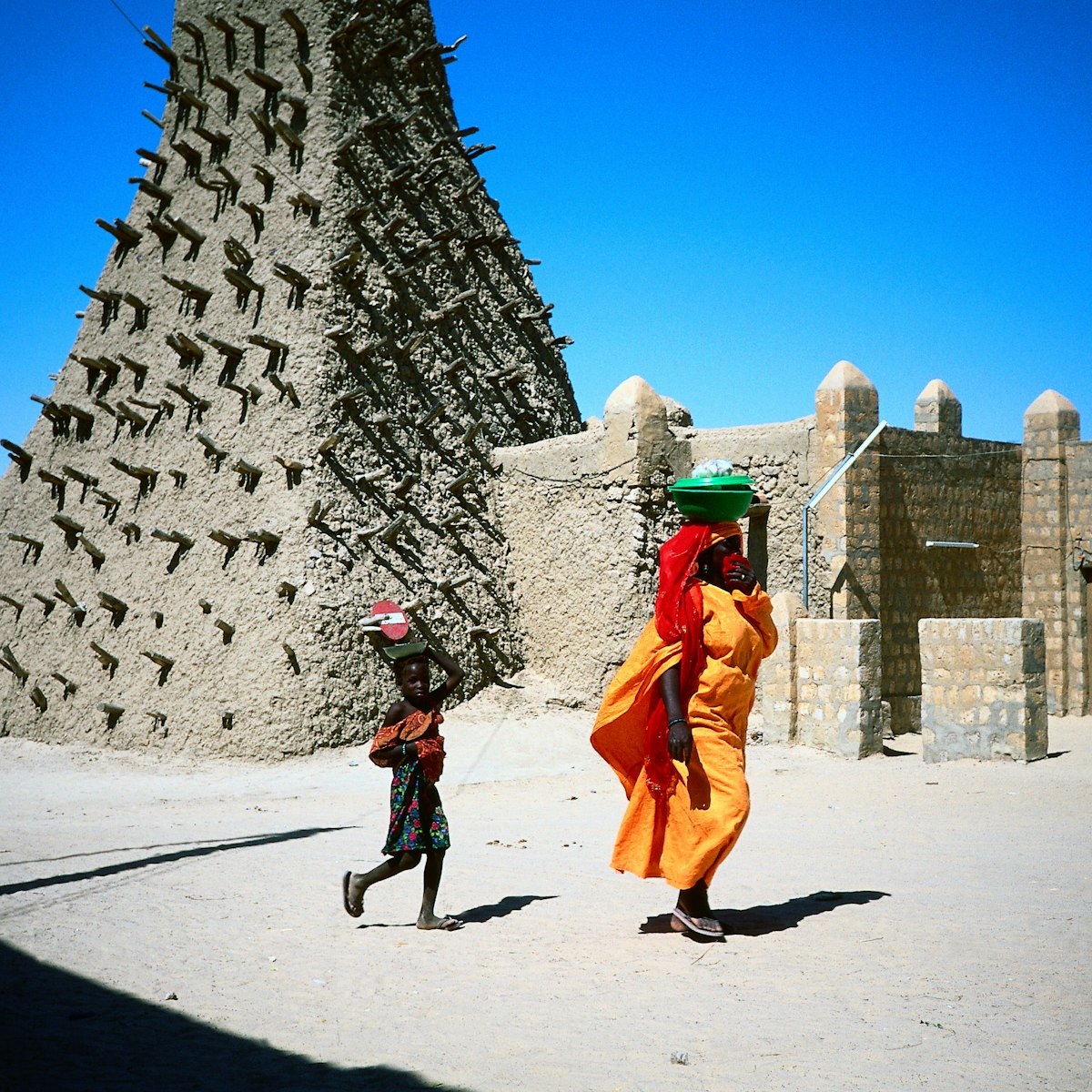 A woman in yellow robes, and a child walk past the Sankore mosque, built in the traditional Sahel style, Timbuktu.