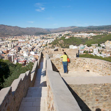 Woman on wall near narrow rampart of Castillo de Gibralfaro, originally built 8th century, looking at view from on high of Malaga, Costa del Sol, Andalucia, Spain