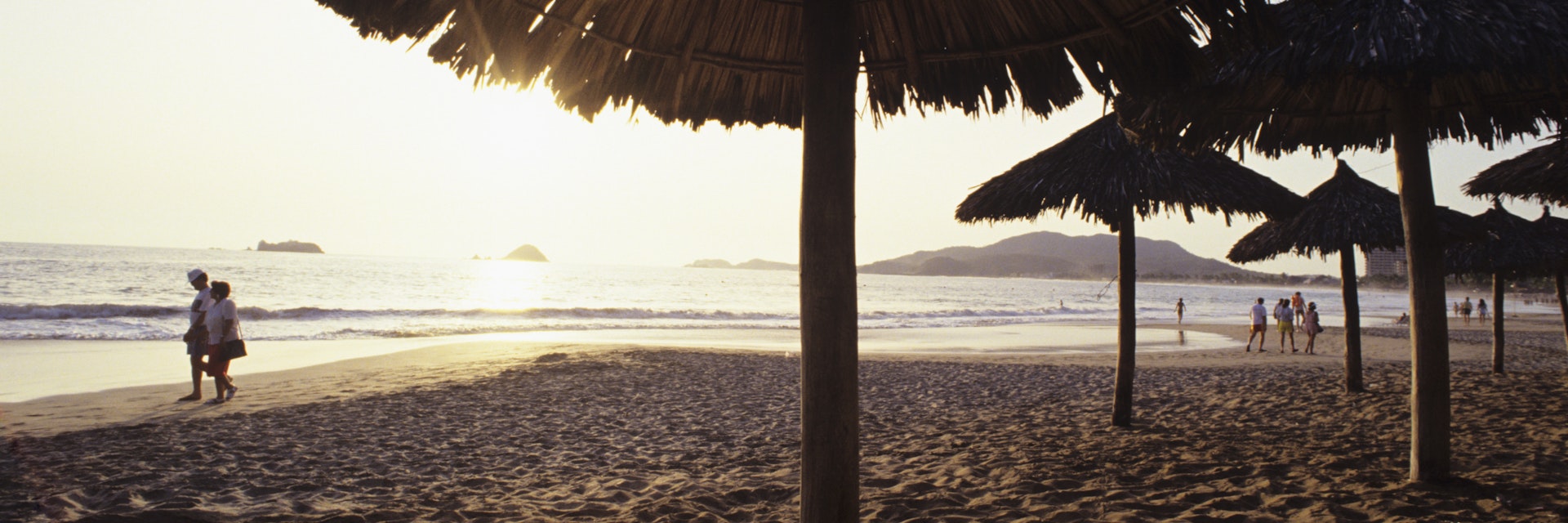 Mexico, Ixtapa, ocean sunset with couples walking along shoreline, umbrellas in sand.