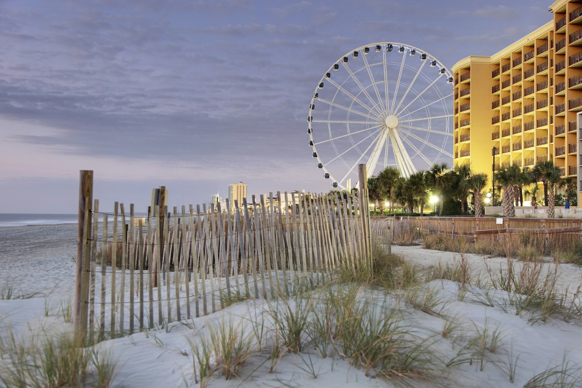 A fence in the dunes along the Boardwalk of Myrtle Beach at dusk, with the SkyWheel and apartment buildings in the distance