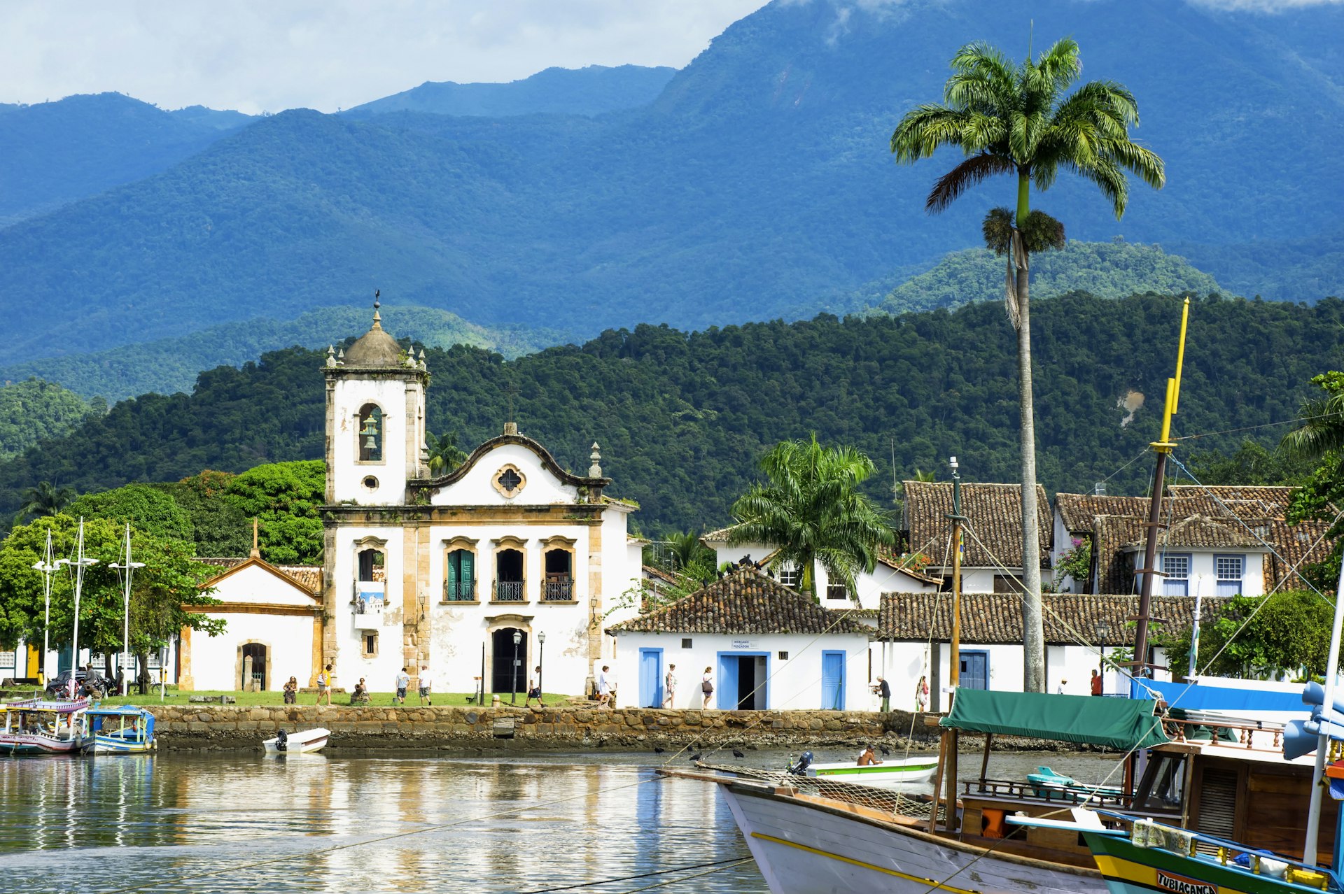 A colonial-era church in Paraty, Brazil, seen from the water, with palm trees and green mountains beyond