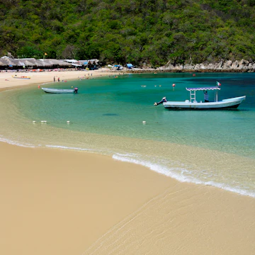 Mexico, Oaxaca, Huatulco, Playa La Entrega, Stretch of sandy beach lined with thatched open fronted restaurants, with tour boat in shallow water in foreground.