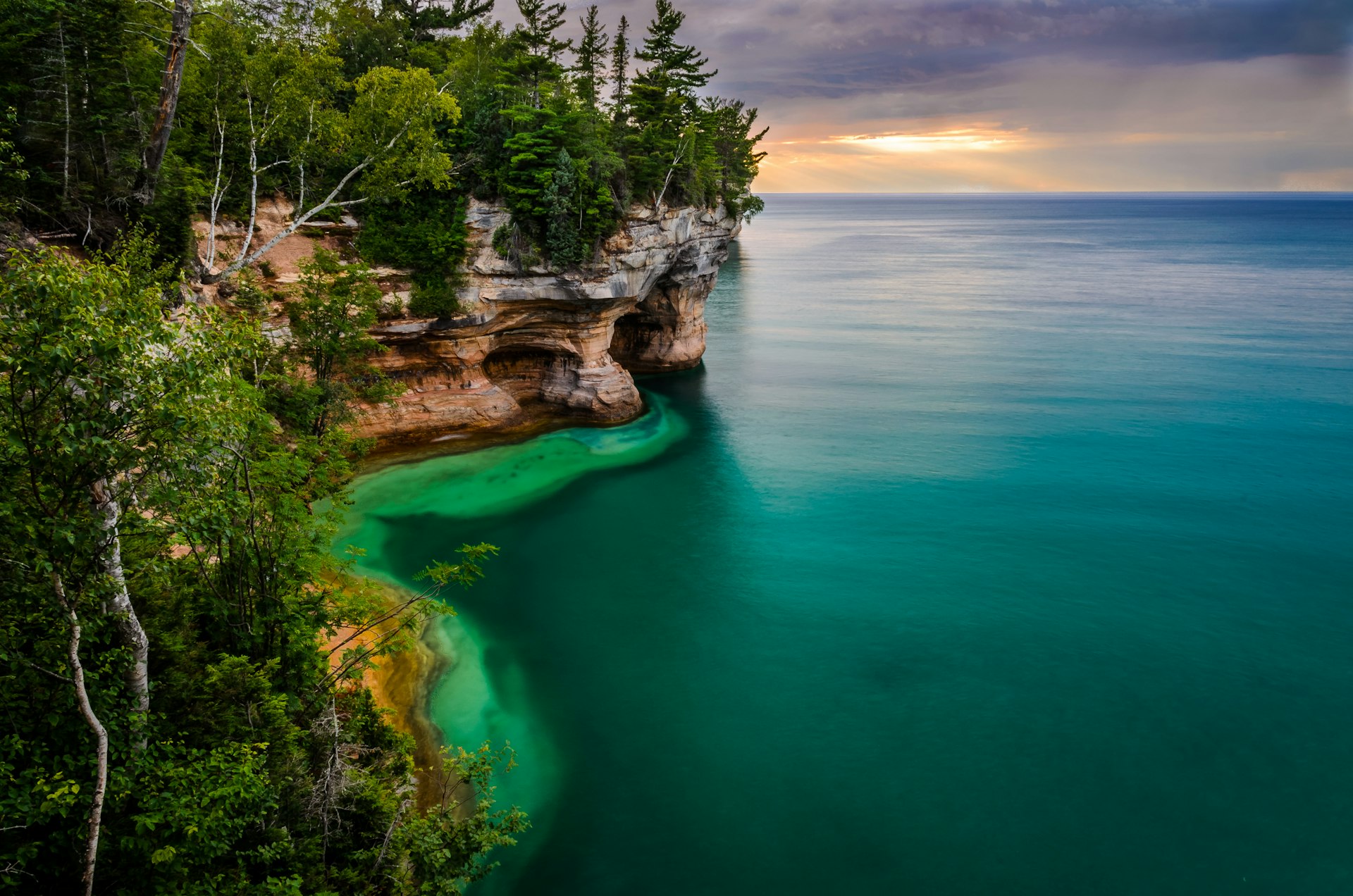 A rocky cliff covered with trees drops into the blue water at Pictured Rocks National Lakeshore in Michigan