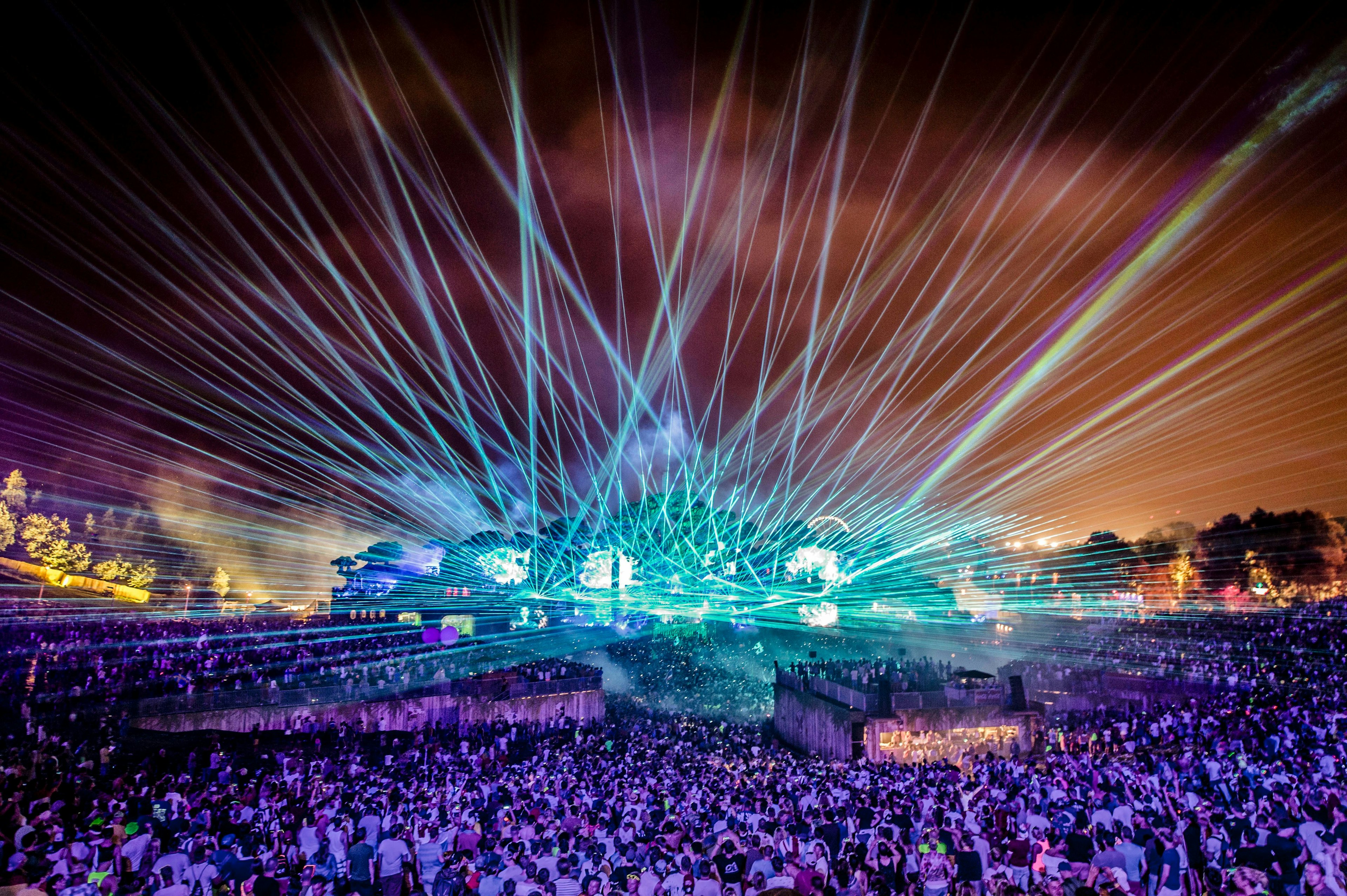 Lasers shoot from a stage into the sky at a music festival with a huge crowd