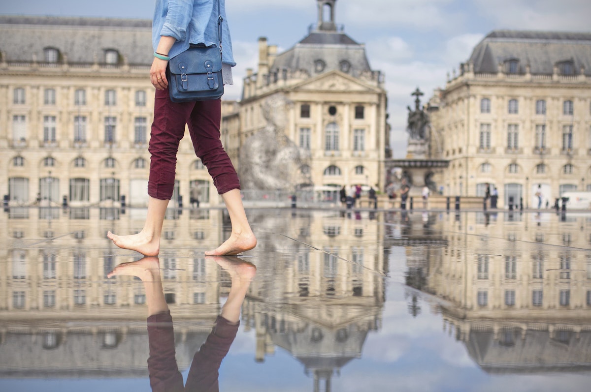 A woman in rolled up burgundy trousers and bare feet, carrying a stylish blue satchel, walks across the Miroir d'Eau water feature at Place de la Bourse in Bordeaux, framed by the historic buildings in the background.