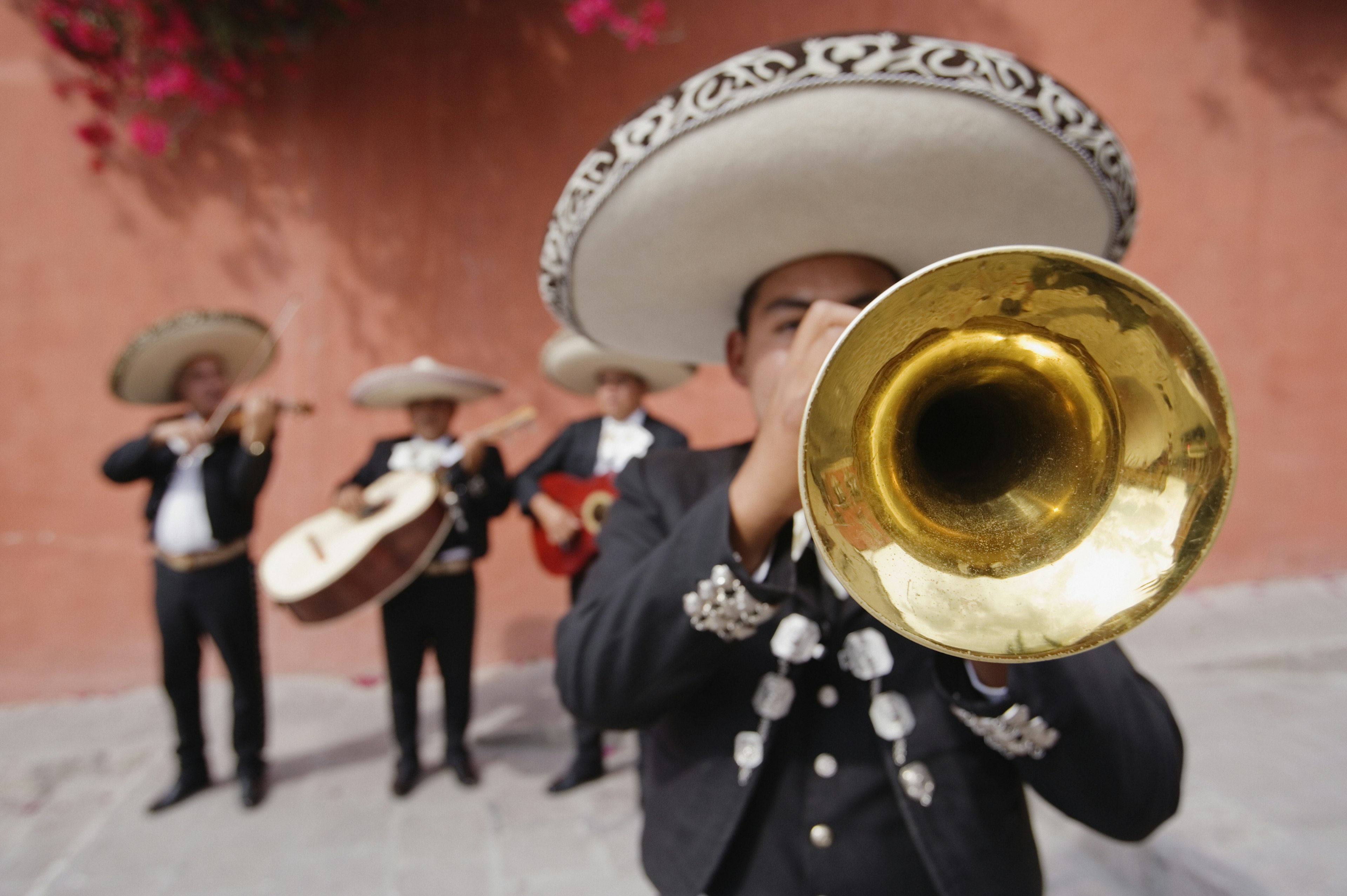 A trumpet player with his face obscured by his instrument and a mariachi band out of focus behind him, all playing in front of a coral-colored wall