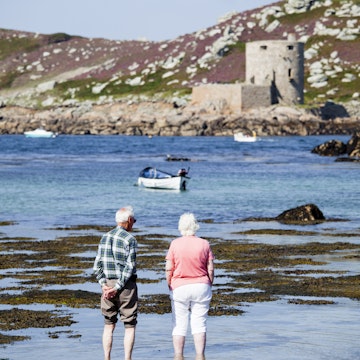 Elderly couple standing in shallows looking towards Cromwell Castle on Tresco Island.