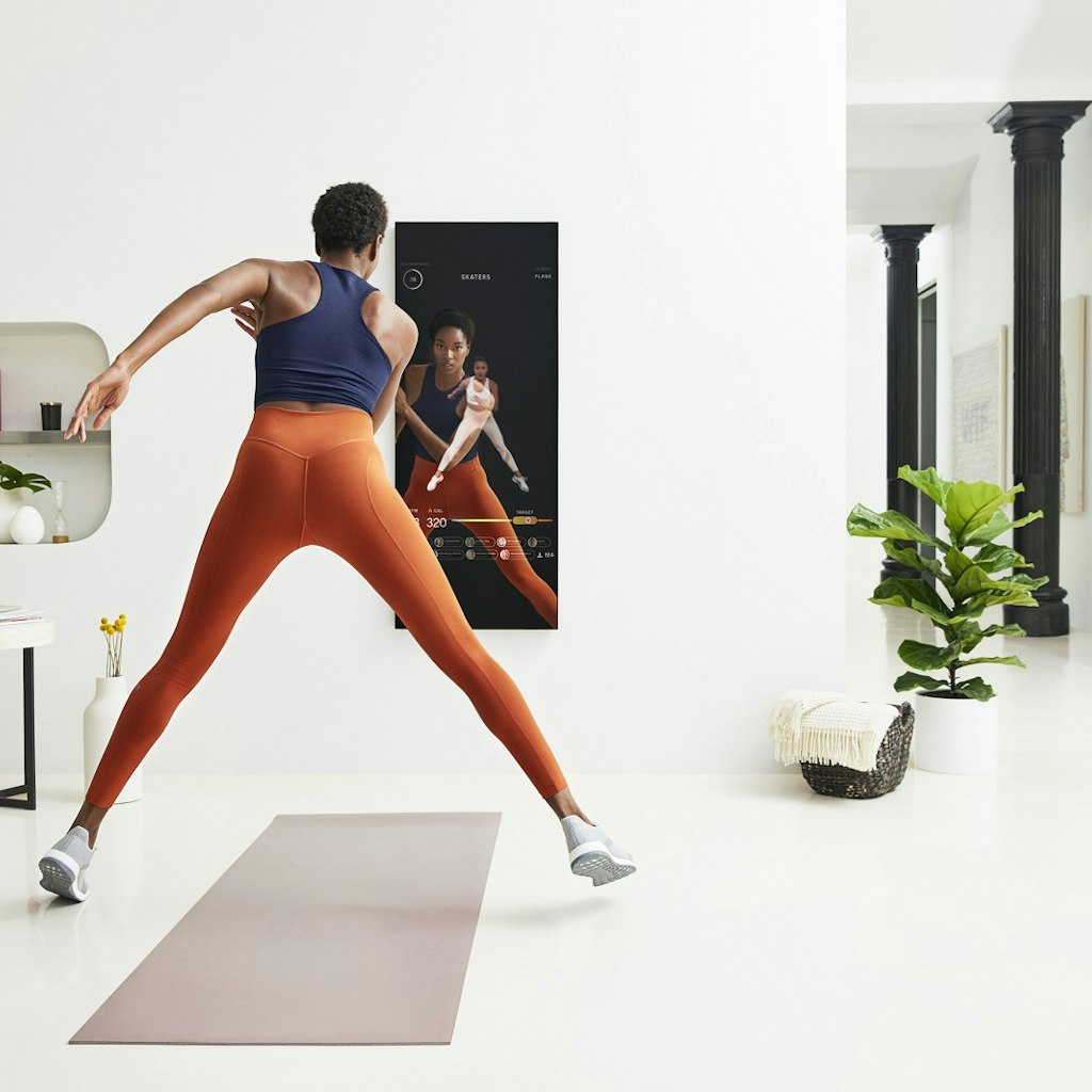 A woman is exercising over a yoga mat and looking at a dark mirror that is instructing her. She is wearing orange leggings and a navy top in a white room that is minimally decorated. 