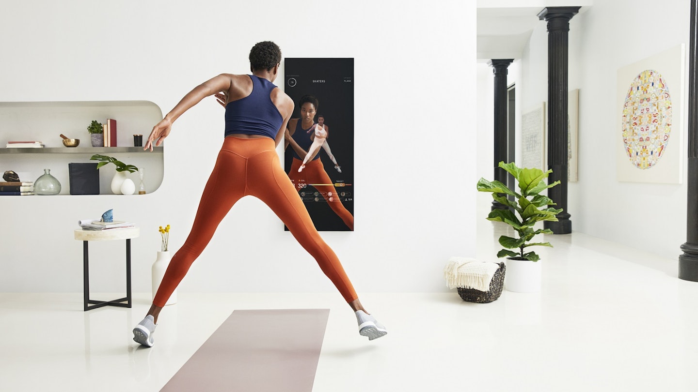 A woman is exercising over a yoga mat and looking at a dark mirror that is instructing her. She is wearing orange leggings and a navy top in a white room that is minimally decorated.