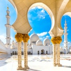 White arches and domes decorated with gold in front of an empty tiled square. Two minarets of the mosque can be seen