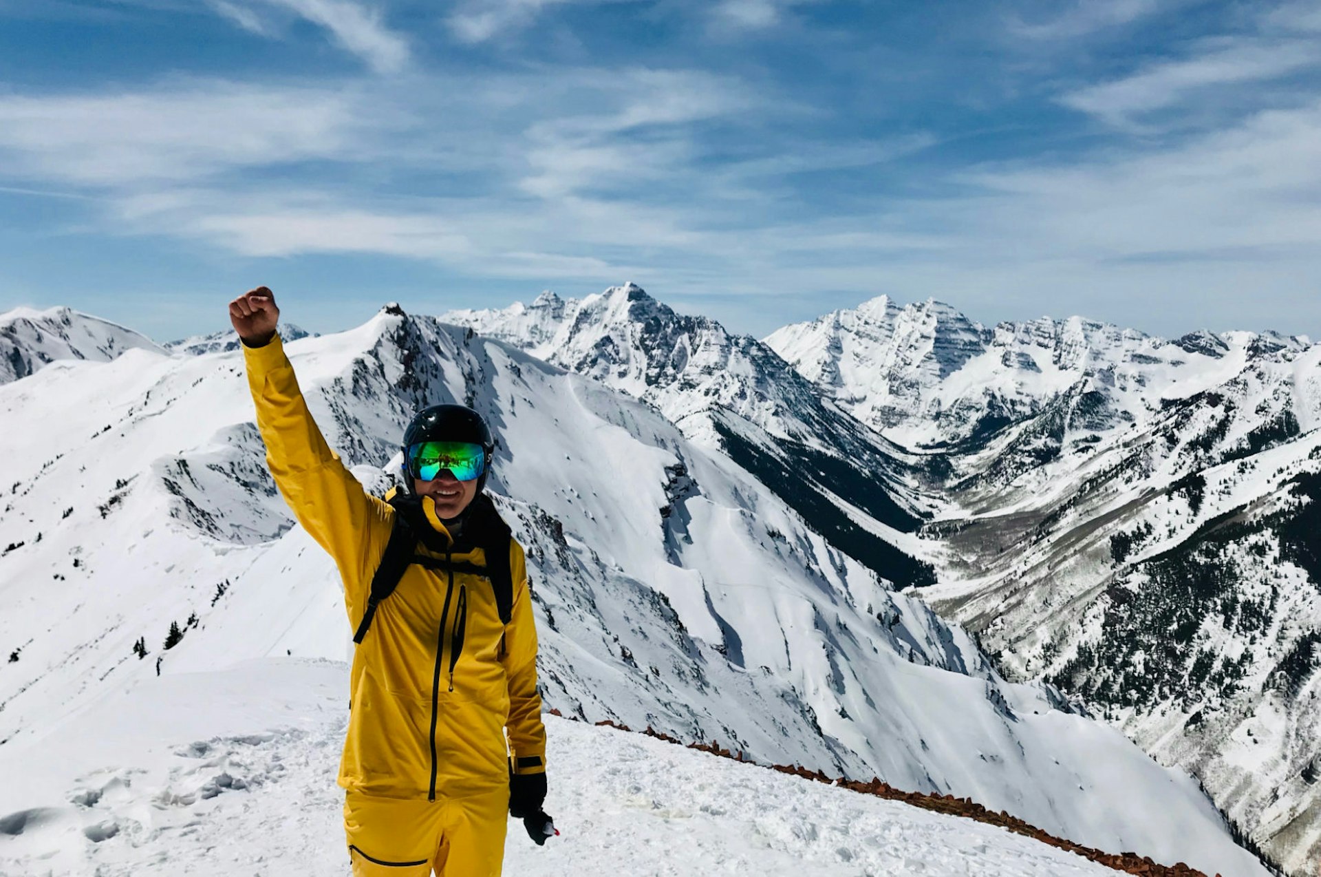 A man stands in front of massifs in Aspen, Colorado wearing the bright yellow waterproof FutureLight pants and jacket, along with goggles, a helmet, and backpack. His right arm is raised in the air with a celebratory fist and he holds his gloves in his left hand by his side.