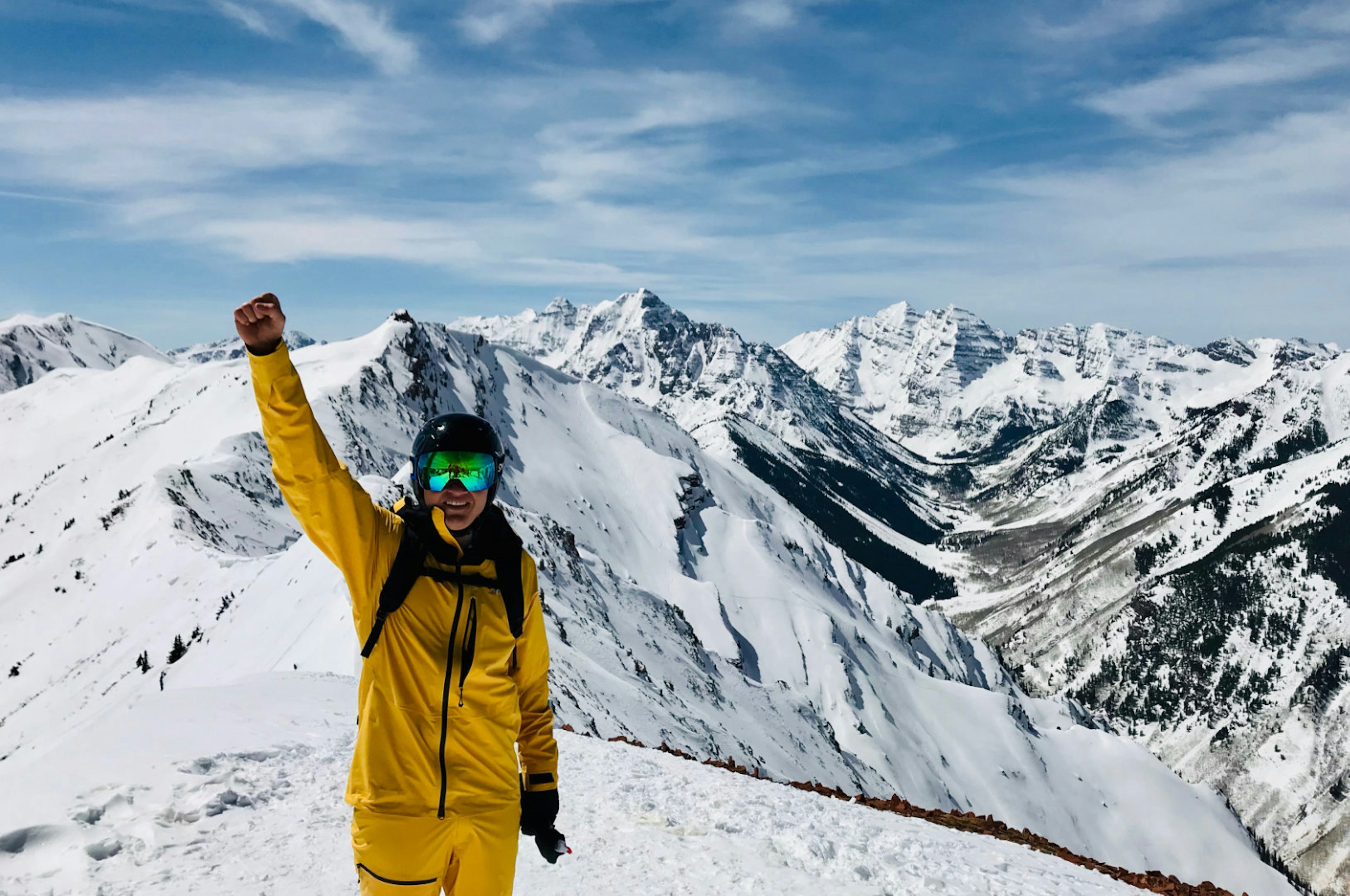Writer Blake Snow stands in front of massifs in Aspen, Colorado wearing the bright yellow waterproof FutureLight pants and jacket, along with goggles, a helmet, and backpack. His right arm is raised in the air with a celebratory fist and he holds his gloves in his left hand by his side.