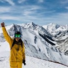 Writer Blake Snow stands in front of massifs in Aspen, Colorado wearing the bright yellow waterproof FutureLight pants and jacket, along with goggles, a helmet, and backpack. His right arm is raised in the air with a celebratory fist and he holds his gloves in his left hand by his side.