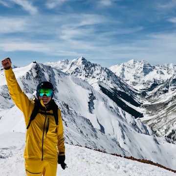 Writer Blake Snow stands in front of massifs in Aspen, Colorado wearing the bright yellow waterproof FutureLight pants and jacket, along with goggles, a helmet, and backpack. His right arm is raised in the air with a celebratory fist and he holds his gloves in his left hand by his side.