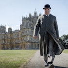 A man in a bowler hat and long beige coat with a stern expression on his face looks directly into the camera as he strides away from Highclere Castle