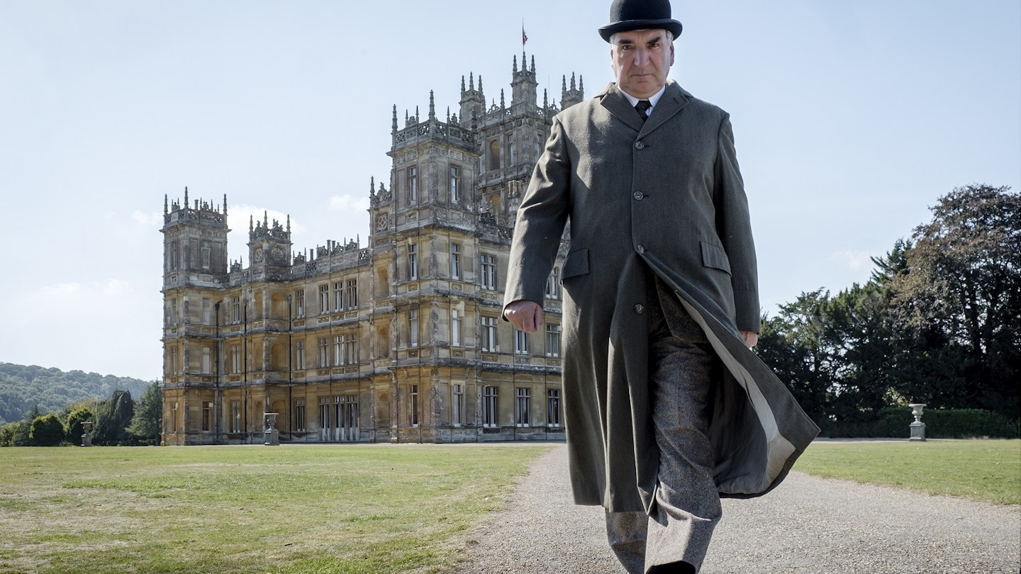A man in a bowler hat and long beige coat with a stern expression on his face looks directly into the camera as he strides away from Highclere Castle