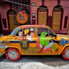 A traditional yellow taxi cab is covered with colorful street art depicting a bright green taxi driver in white kurti pants with a red Kartrī sword and a grey turban. On the rear door is a lion, with the faces of passengers painted over the window. Behind the taxi is a pink stucco house with brown shutters. 