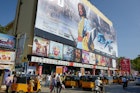 Yellow motorized rickshaws and pedestrians move down the street in front of a movie theater in Chennai plastered with movie posters, including a large multi-story ad featuring a male movie star with long hair and traditional Indian costume.