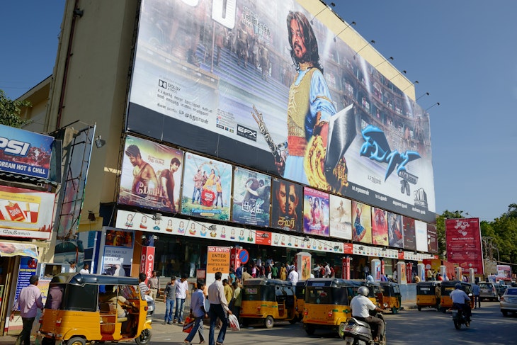 Yellow motorized rickshaws and pedestrians move down the street in front of a movie theater in Chennai plastered with movie posters, including a large multi-story ad featuring a male movie star with long hair and traditional Indian costume.