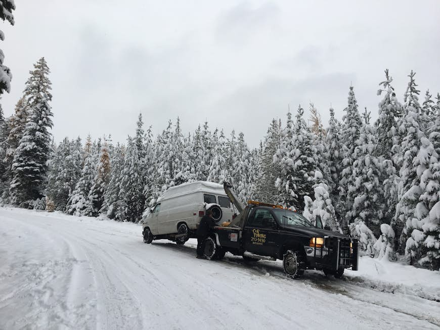 En vit GMC skåpbil bogseras av en svart vrakbil på en snöig väg i Montana.  Granar och tallar i bakgrunden vid snötäckta, liksom skogsvägen där skåpbilen gick sönder.