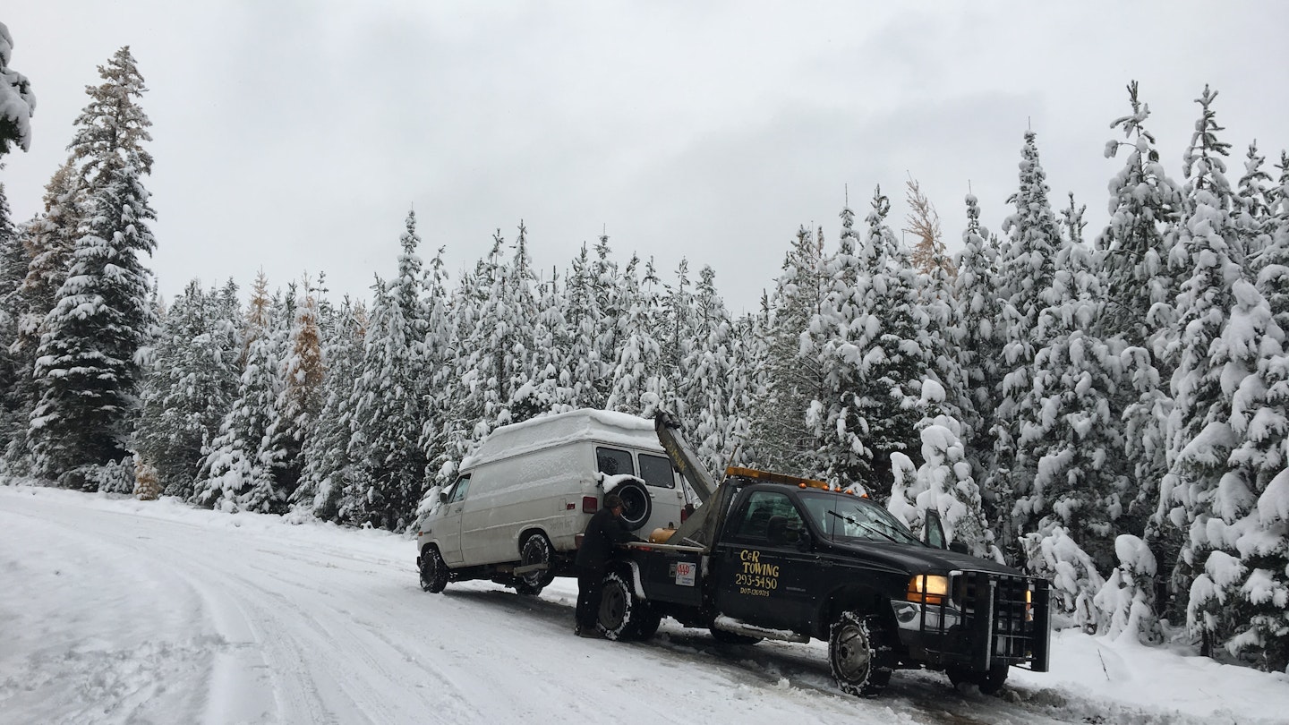 A white GMC van is being towed by a black wrecker on a snowy road in Montana. Spruce and pine trees in the background at blanketed with snow, as is the forest service road where the van broke down.