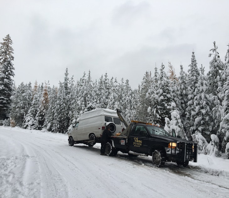 A white GMC van is being towed by a black wrecker on a snowy road in Montana. Spruce and pine trees in the background at blanketed with snow, as is the forest service road where the van broke down.