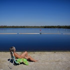 A nude, middle-aged woman with short grey hair lounges in a beach chair on a lime green towel with her feet in the sand in front of a deep blue lake reflecting the sky. A think band of short green trees lines the rim of the lake in the background. 