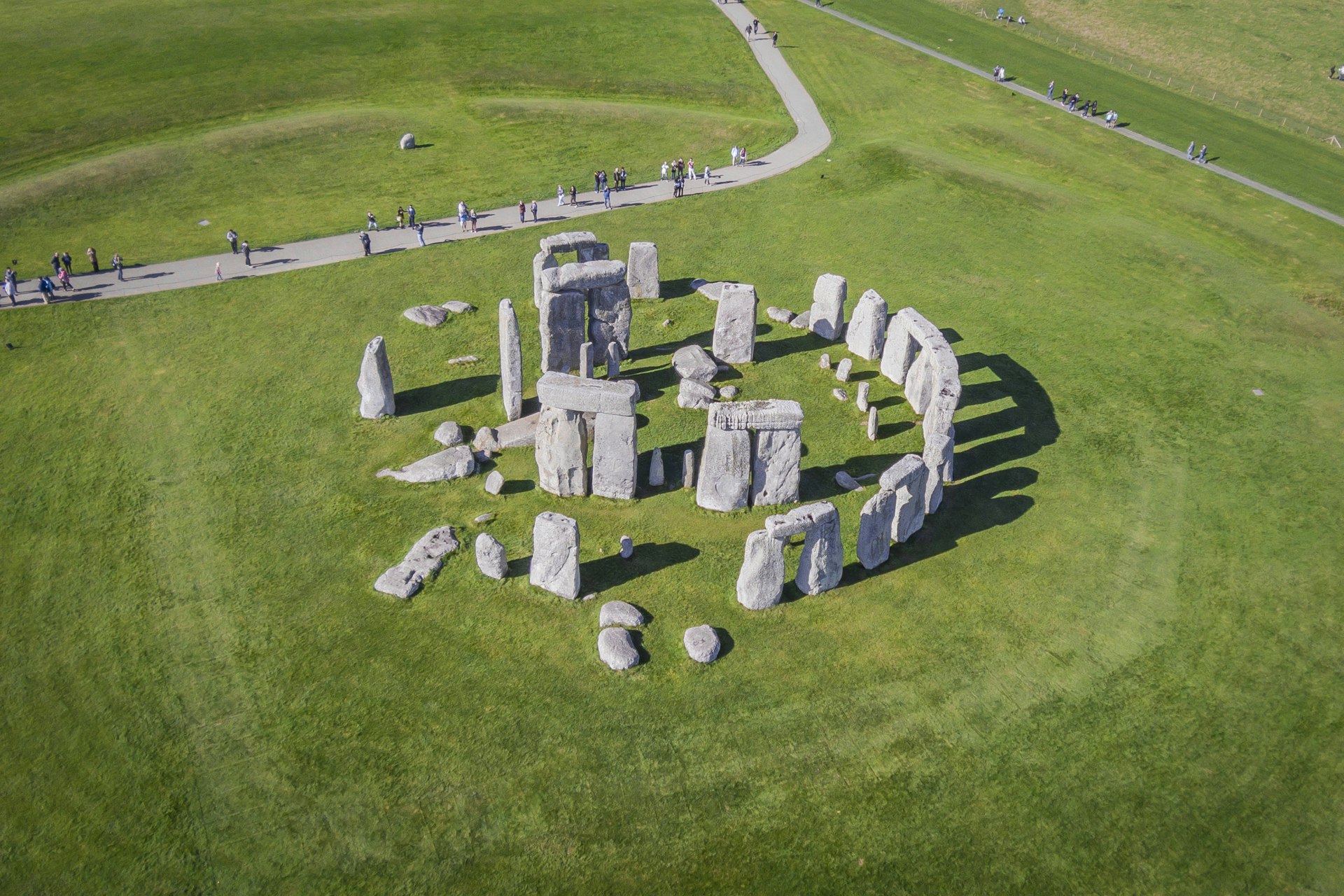 Aerial shot of Stonehenge, a collection of giant grey stones arranged in two circles. There are some visitors at the site, who seem tiny when compared to the size of the stones