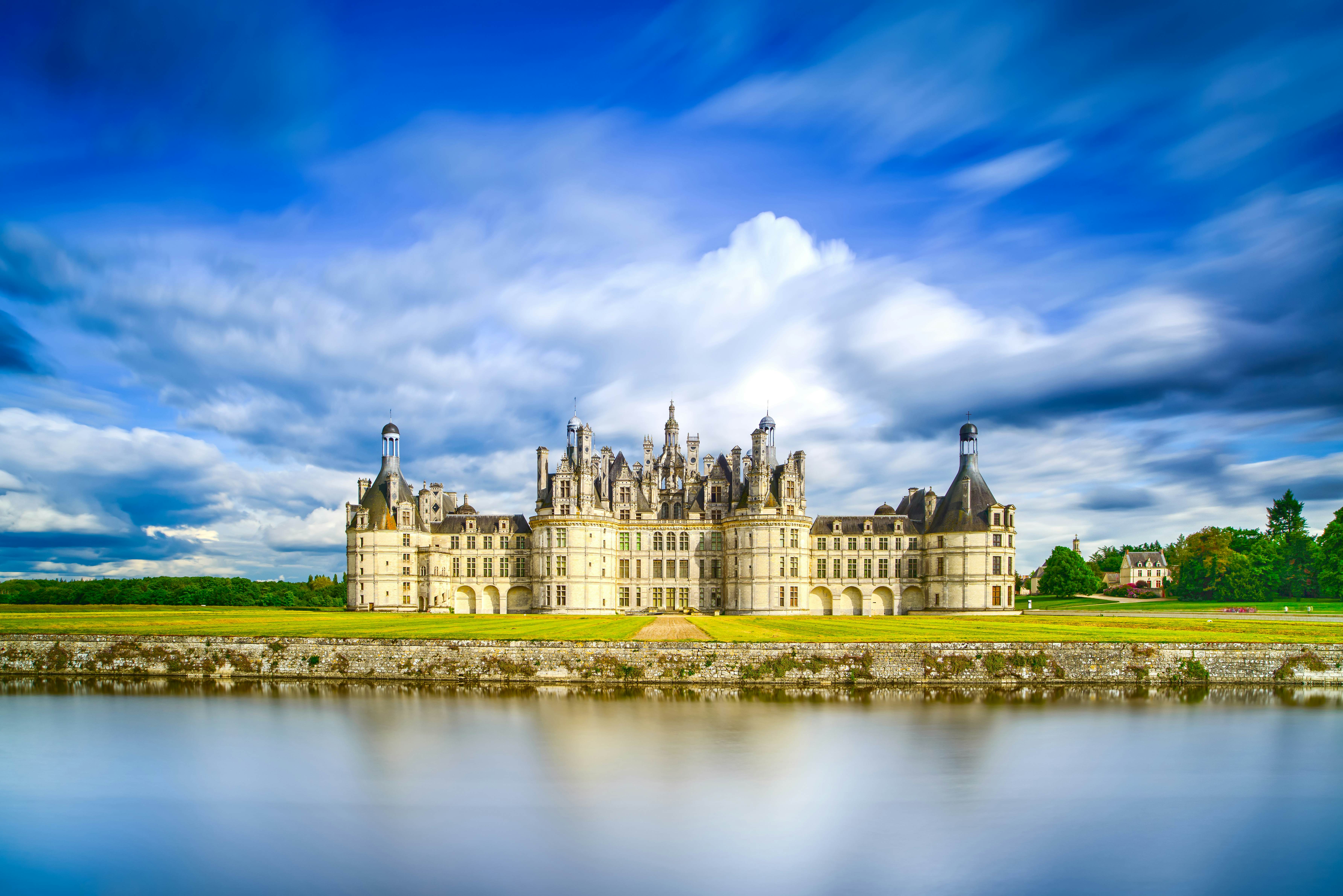 A large white chateau with several huge turrets photographed from a distance and reflected in a river