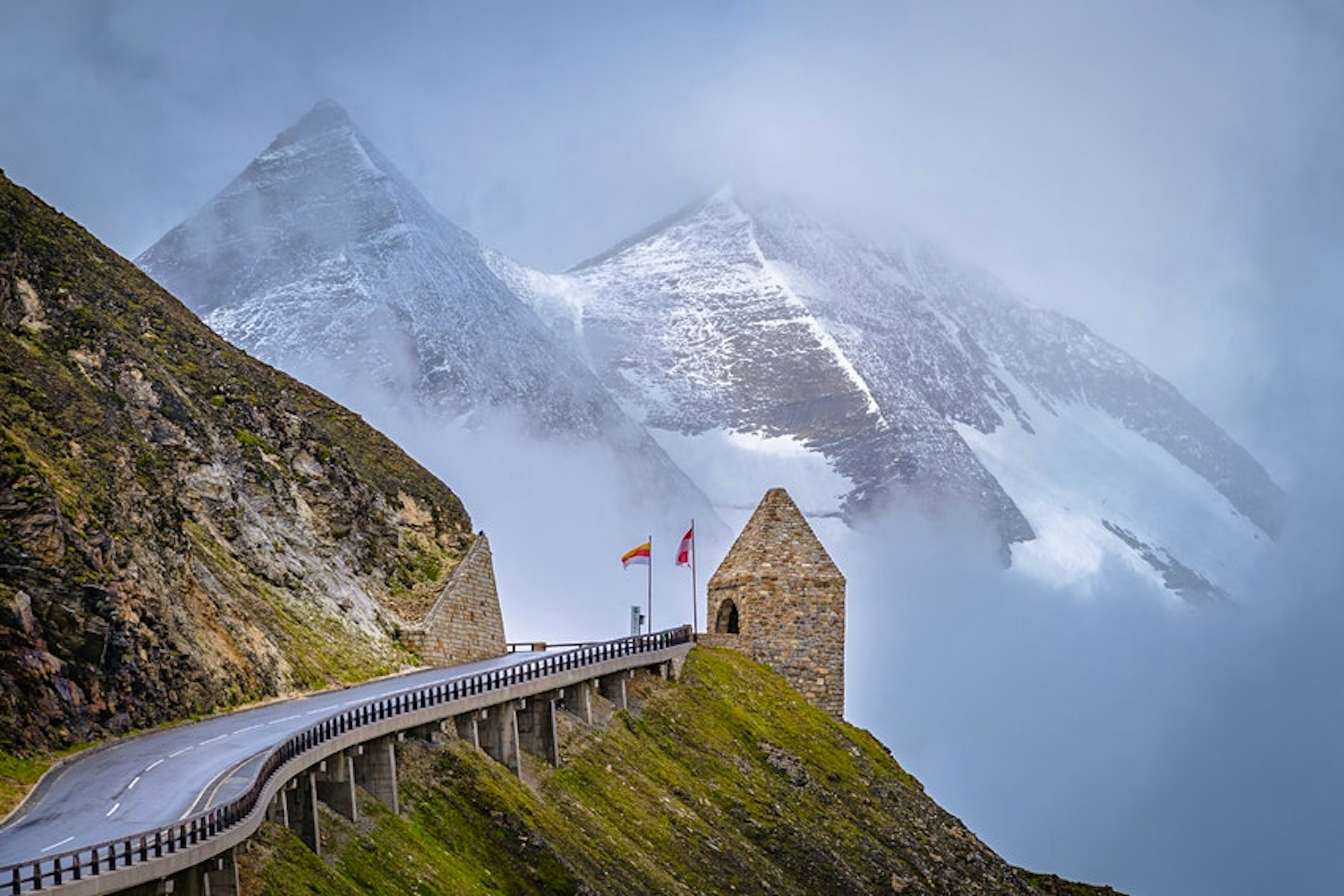 A two-lane highway along tall snow-capped mountains. There is a stone structure coming out of the side of a mountain with two flags in front of it