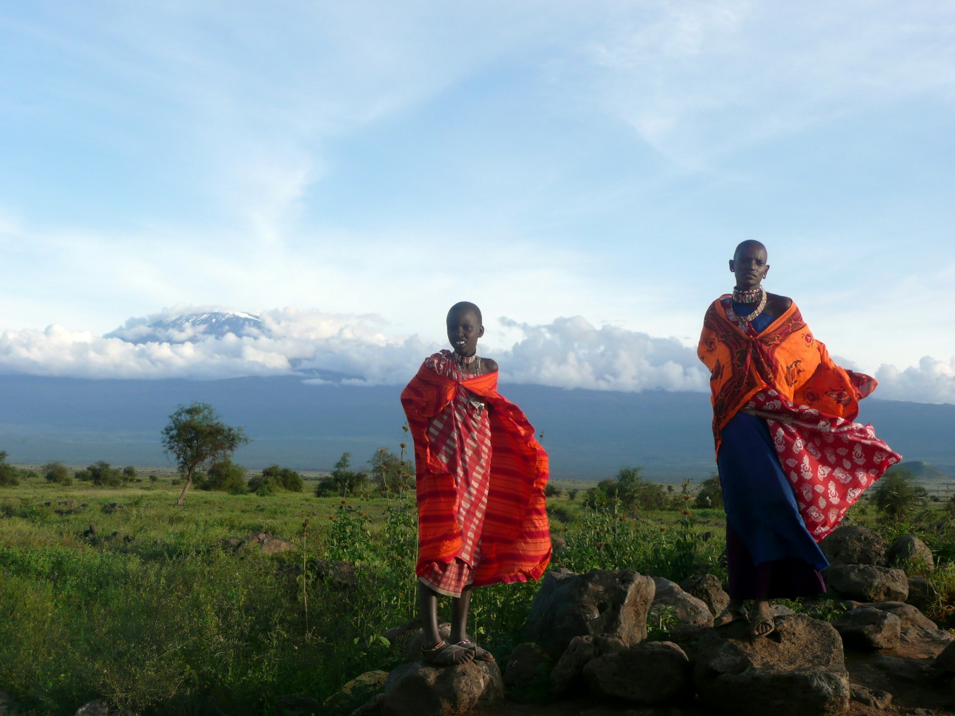 A pair of Maasai dressed in brightly colored garb stand on a pair of tall rocks. In the background, you can see Mt. Kilimanjaro covered by dense clouds in the distance. 