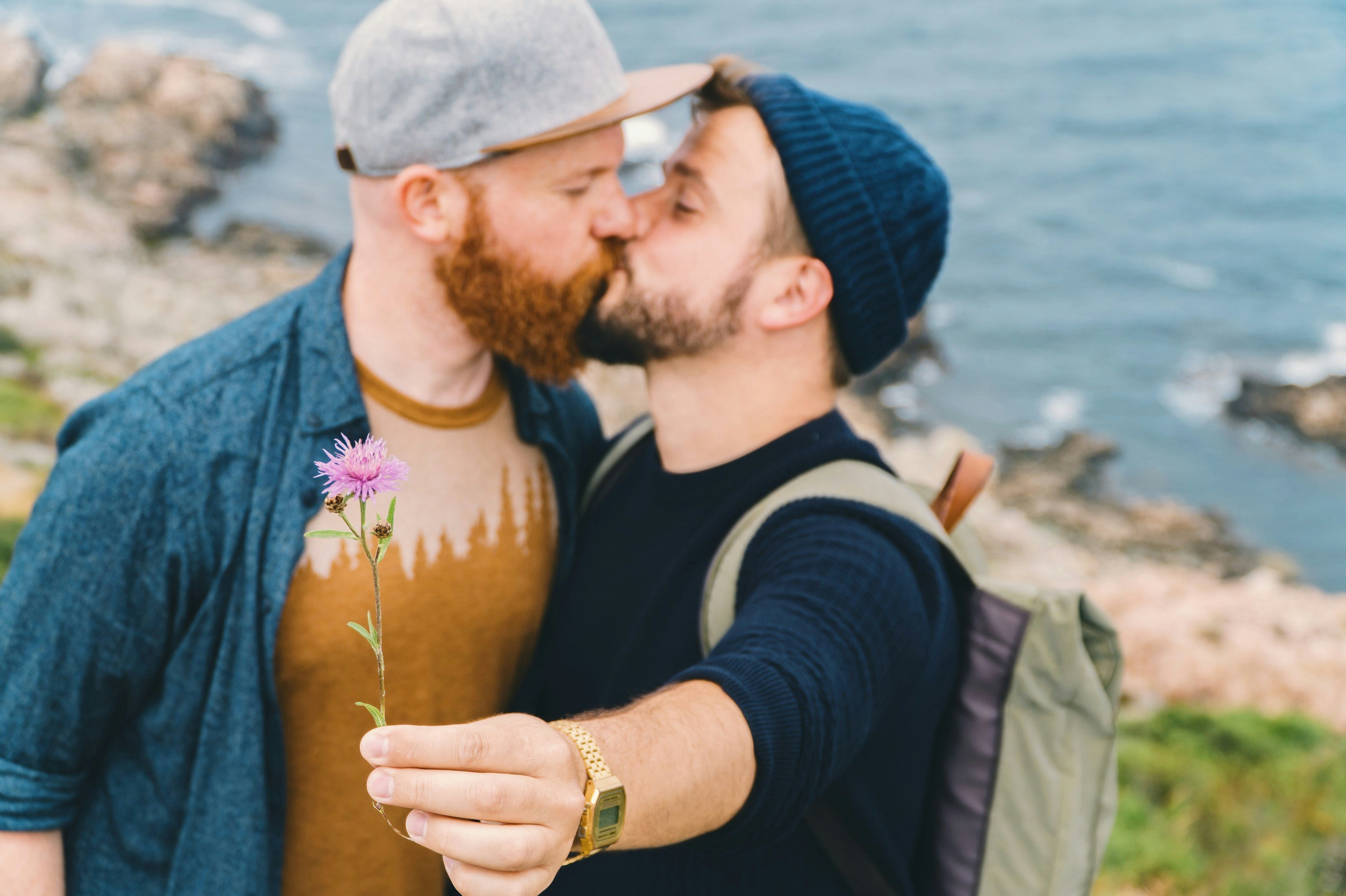 Two white men kissing on a clifftop. They're slightly out of focus, with the camera zoned in on a small purple flower that one man is holding out