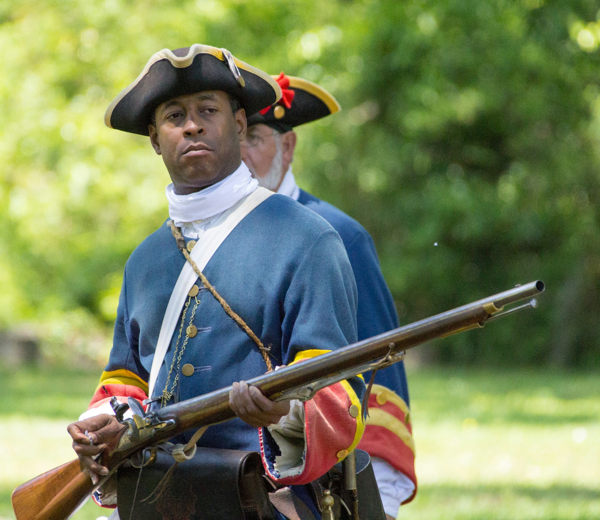 A black man dressed in Colonial solider garb holds a wooden musket 