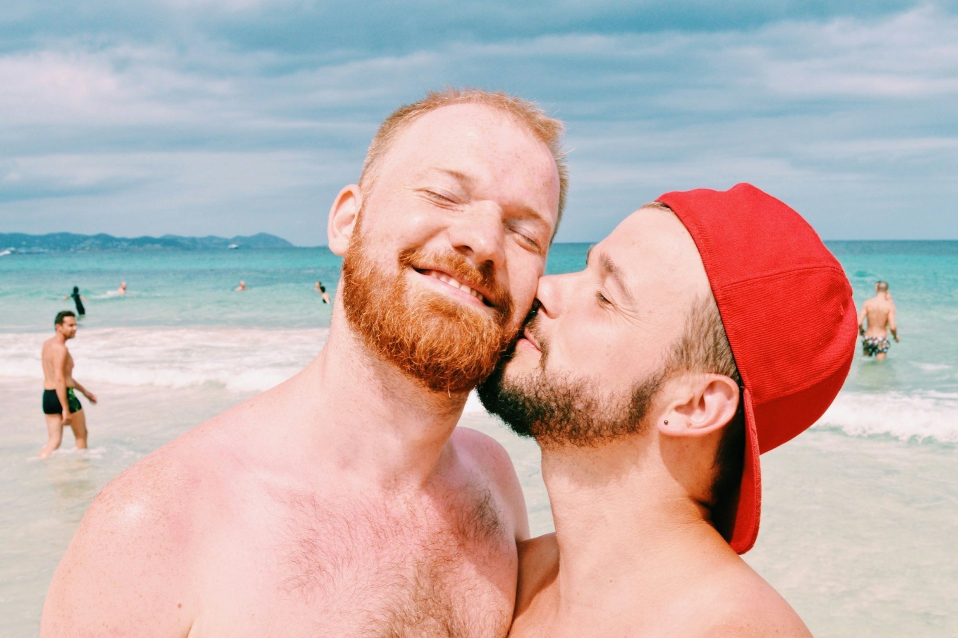 Two white men with beards on a beach. One is wearing a red cap backwards and is kissing the other man on the cheek
