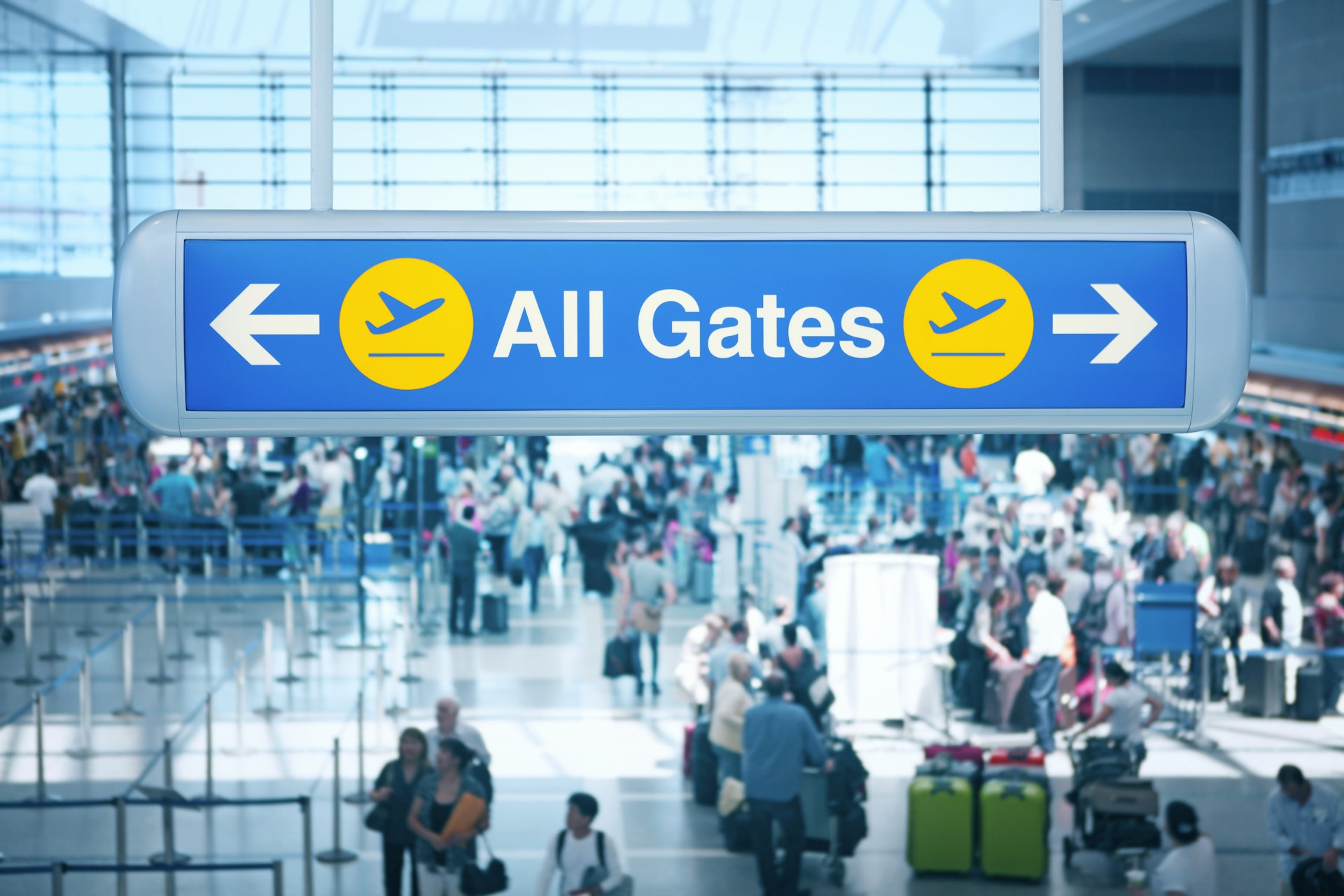 A large sign with "All Gates" written on it at an airport. Groups of people with luggage are lining up at desks in the concourse below the sign