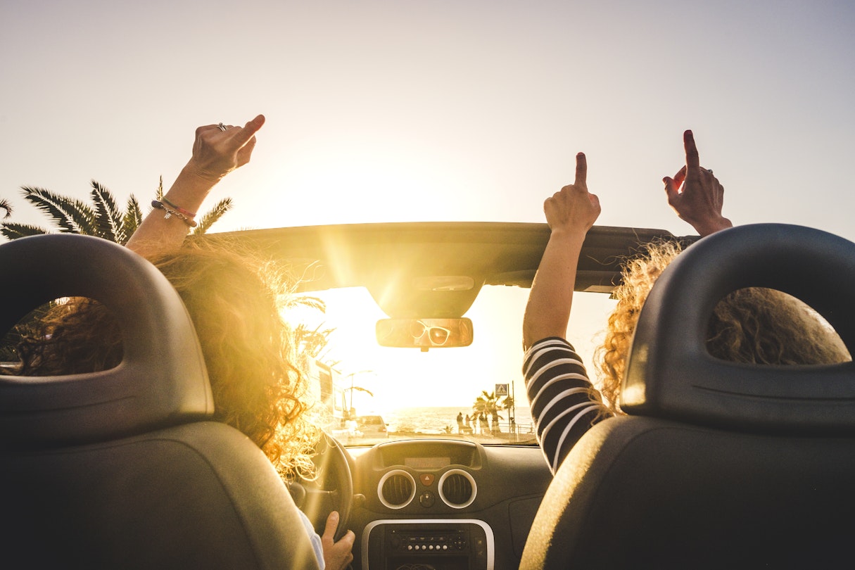 Couple of female friends driving in a convertible and raising their hands to music with the sun setting over the ocean.