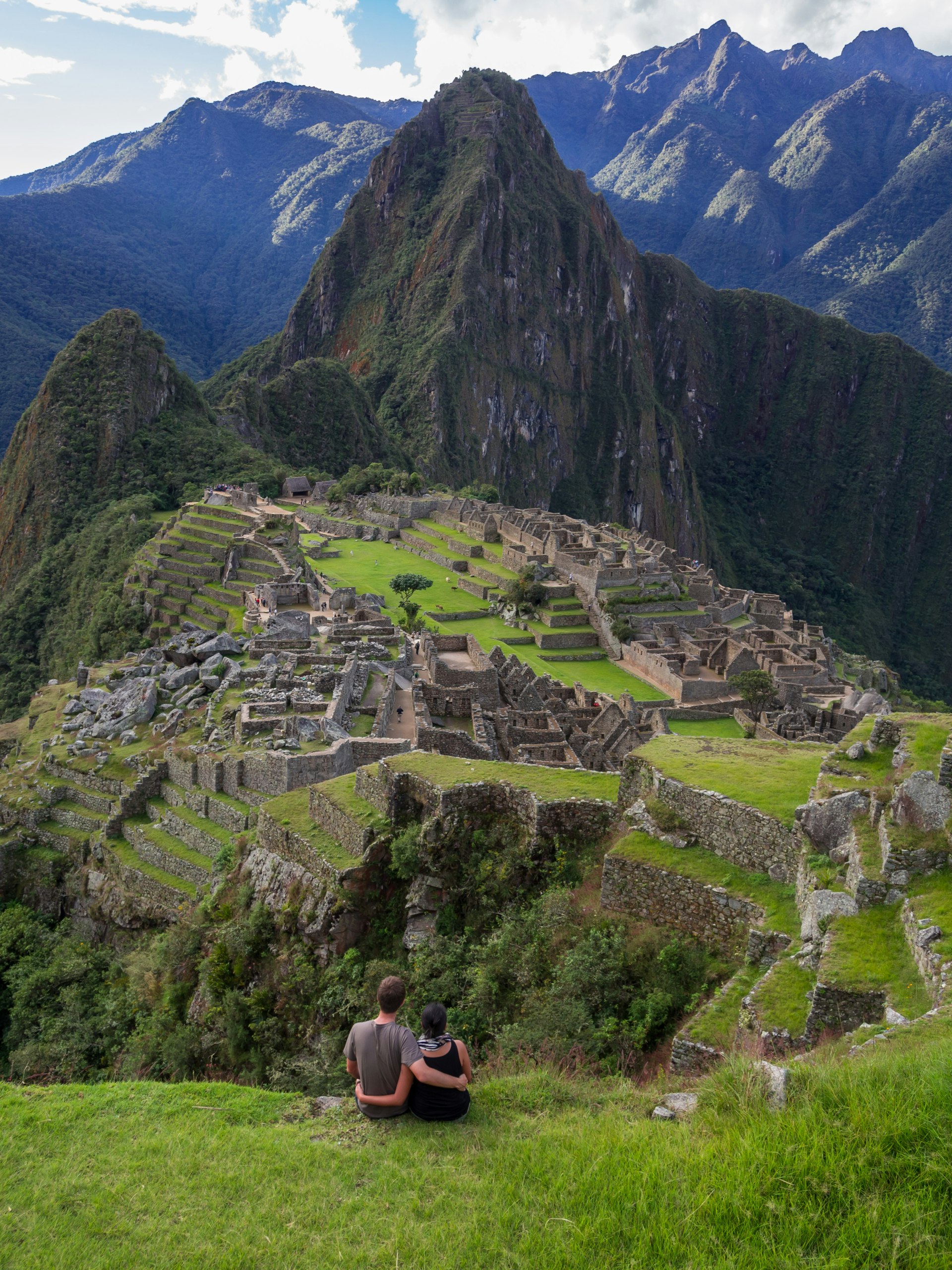 A couple sits on a grassy verge with their arms around each other's waists, looking down at Machu Picchu.