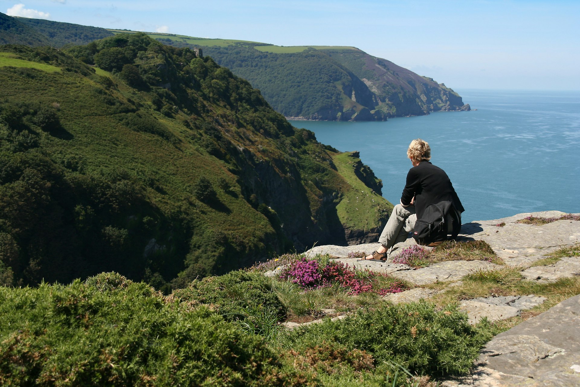 A solo figure sits on a clifftop looking along a coastline that stretches off into the distance