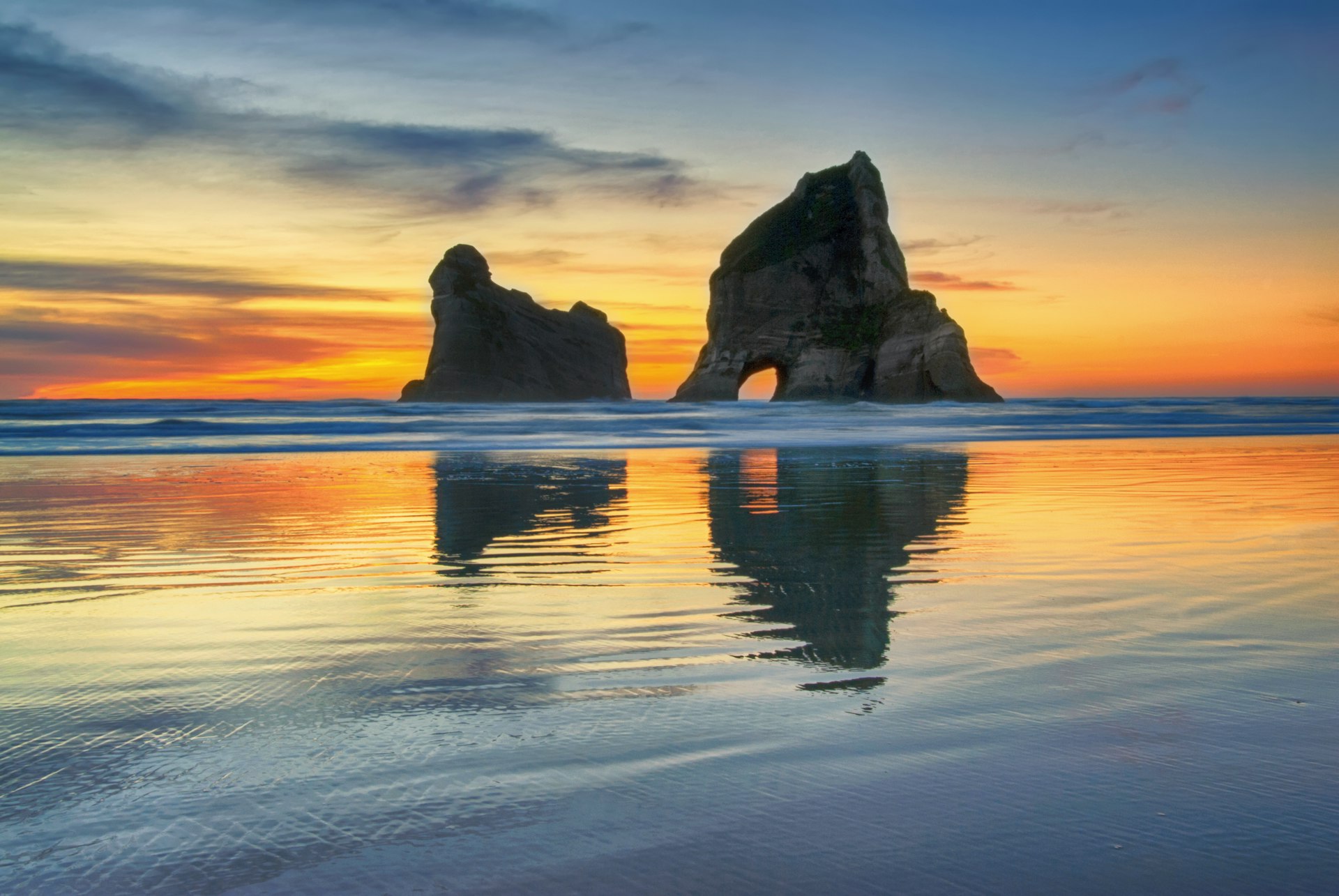 A red band stretches across the horizon as the sun sets on stone archways at Wharariki Beach in New Zealand 