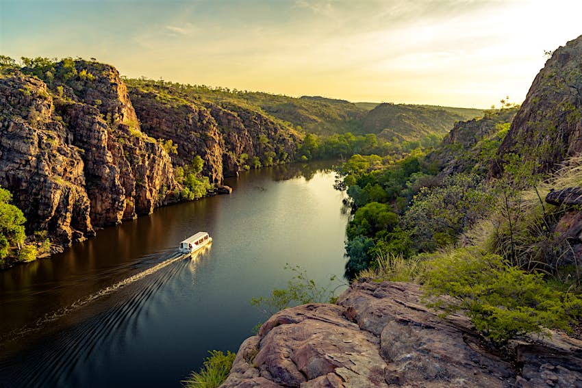 A white boat sails down Katherine Gorge and is flanked by jagged rocks covered in lush green trees and shrubs